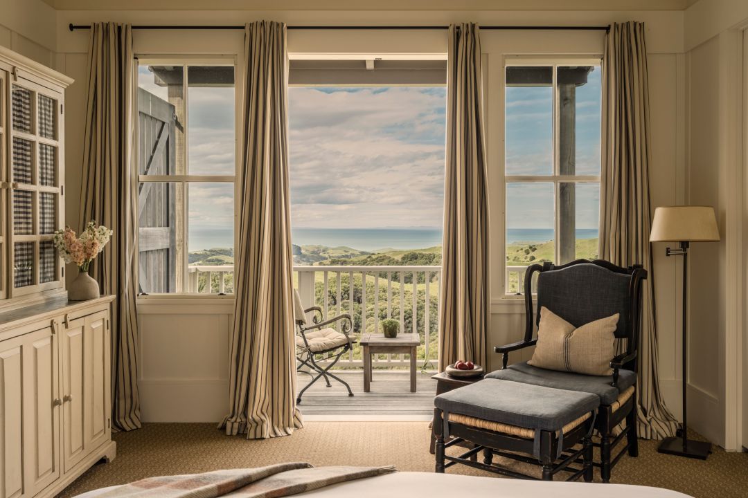 A hotel suite with neutral furnishings including a plush bed, chair and balcony with ocean views. 